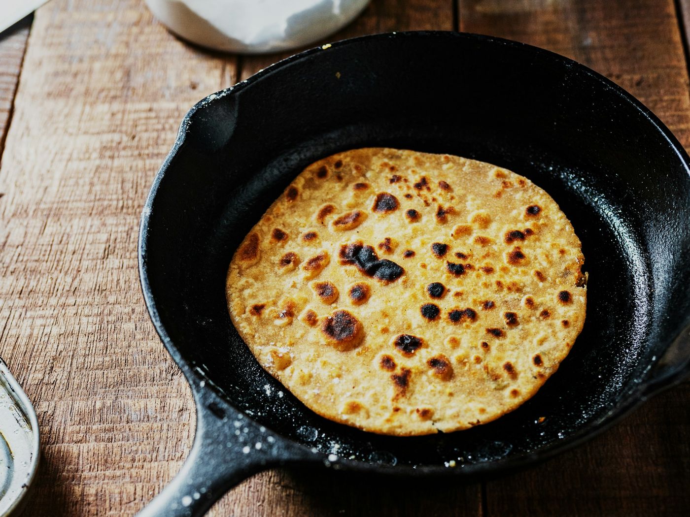 A cast iron skillet with a golden, lightly charred flatbread on a rustic wooden table dusted with flour.